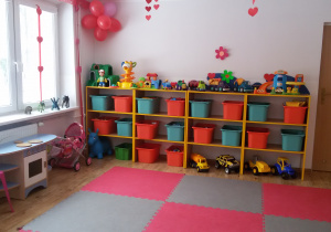 Widok na salę przedszkolną, na wprost znajduje się regał z zielonymi i pomarańczowymi pojemnikami, na górze stoją zabawki, po lewej stronie, pod oknem stoi kącik kuchenny, różowy plastikowy stolik i wózek dla lalek.
